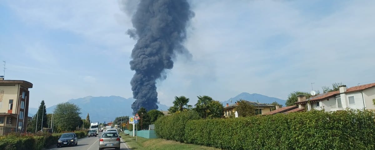 Colonna di fumo scuro dell’incendio di Malo oltre una strada, con case, alberi e rilievi sullo sfondo