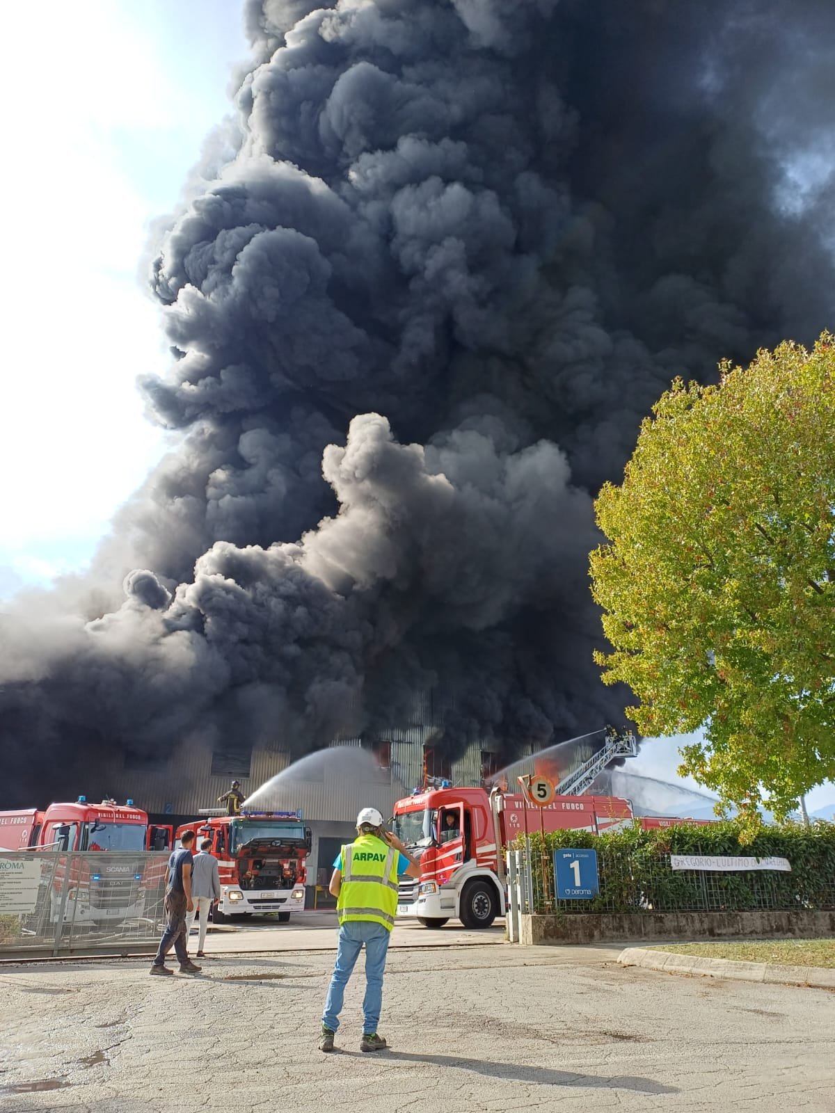 Vigili del fuoco che dirigono getti d’acqua sul capannone di Malo sotto una colonna di fumo scuro; in primo piano un tecnico ARPAV